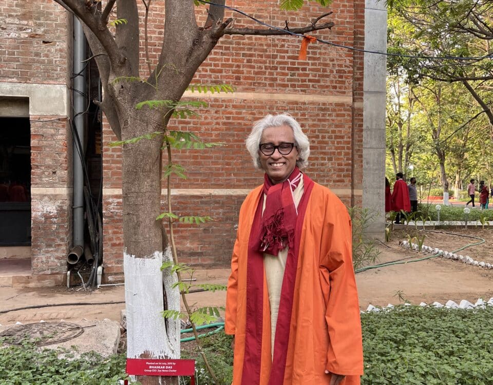 Man in vibrant outfit smiles warmly in front of a brick building and tree, embodying the legacy of Bhaskar Das: Media Pioneer.
