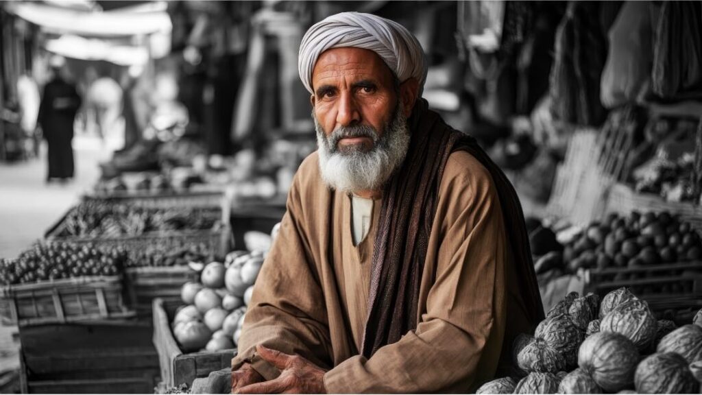 An elderly man with a white beard and turban sits at a market stall surrounded by crates of fresh produce in a black and white photograph.